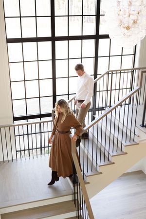 Man and woman descending modern staircase with black railings, large windows, and chandelier in elegant home.