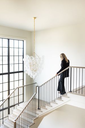 Woman in black outfit standing on modern staircase with elegant bubble chandelier and large windows in bright interior.