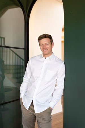 Smiling man in white button-up shirt and khaki pants standing in green arched doorway of modern home.
