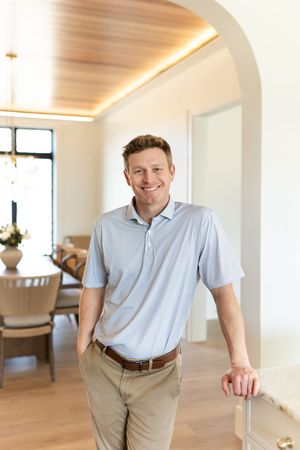 Smiling man in light blue polo shirt leaning on staircase railing in modern home interior.