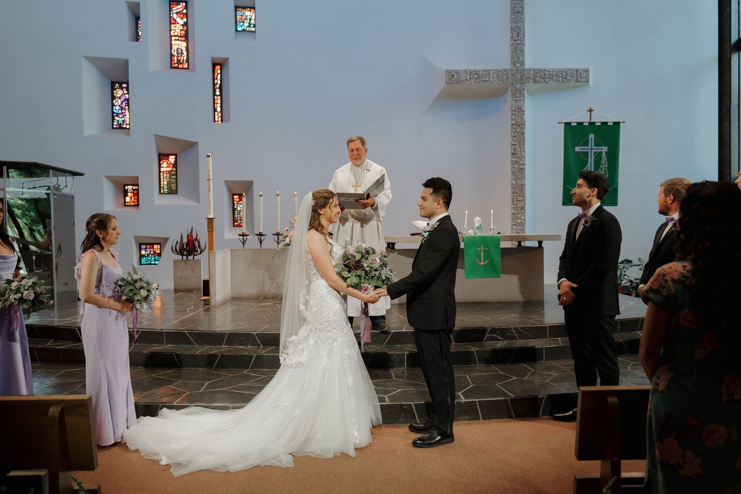 Bride and groom exchange vows at church altar with officiant, bridesmaid, and groomsman during wedding ceremony.