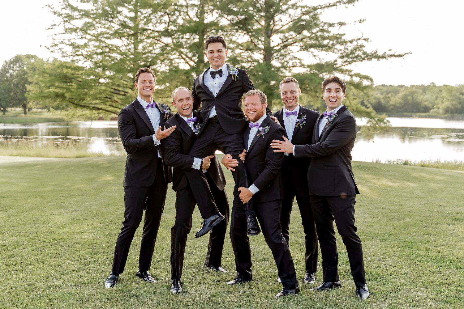 Groomsmen in black tuxedos lift groom outdoors near scenic pond and trees during wedding celebration.