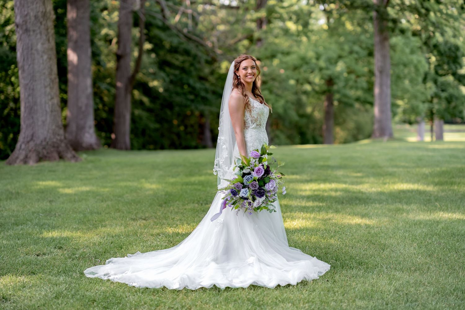 Bride in elegant white gown with long train holds purple bouquet on lush green lawn surrounded by tall trees.