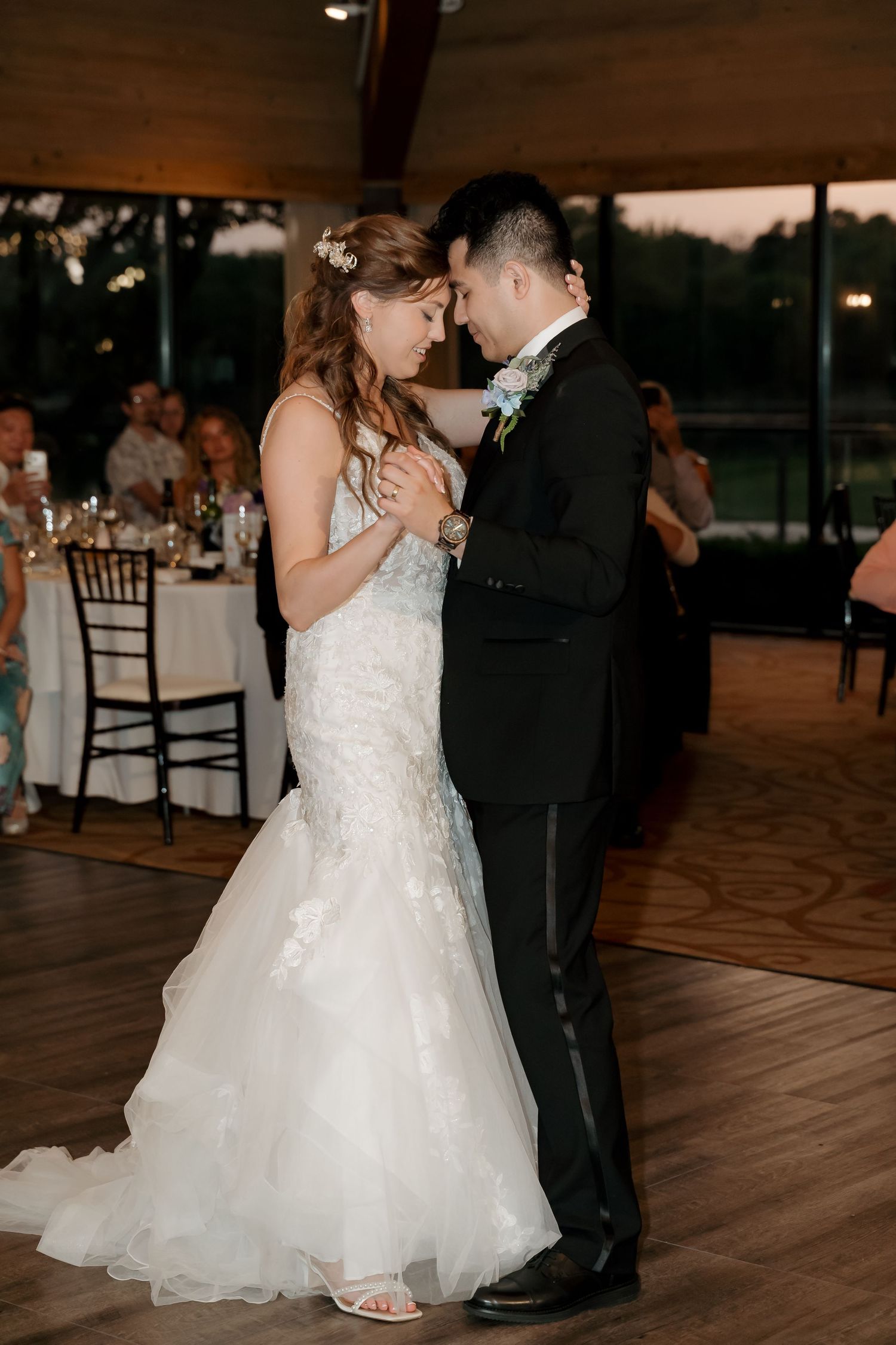 Bride and groom share their first dance at wedding reception, bride in lace mermaid gown, groom in black tuxedo.