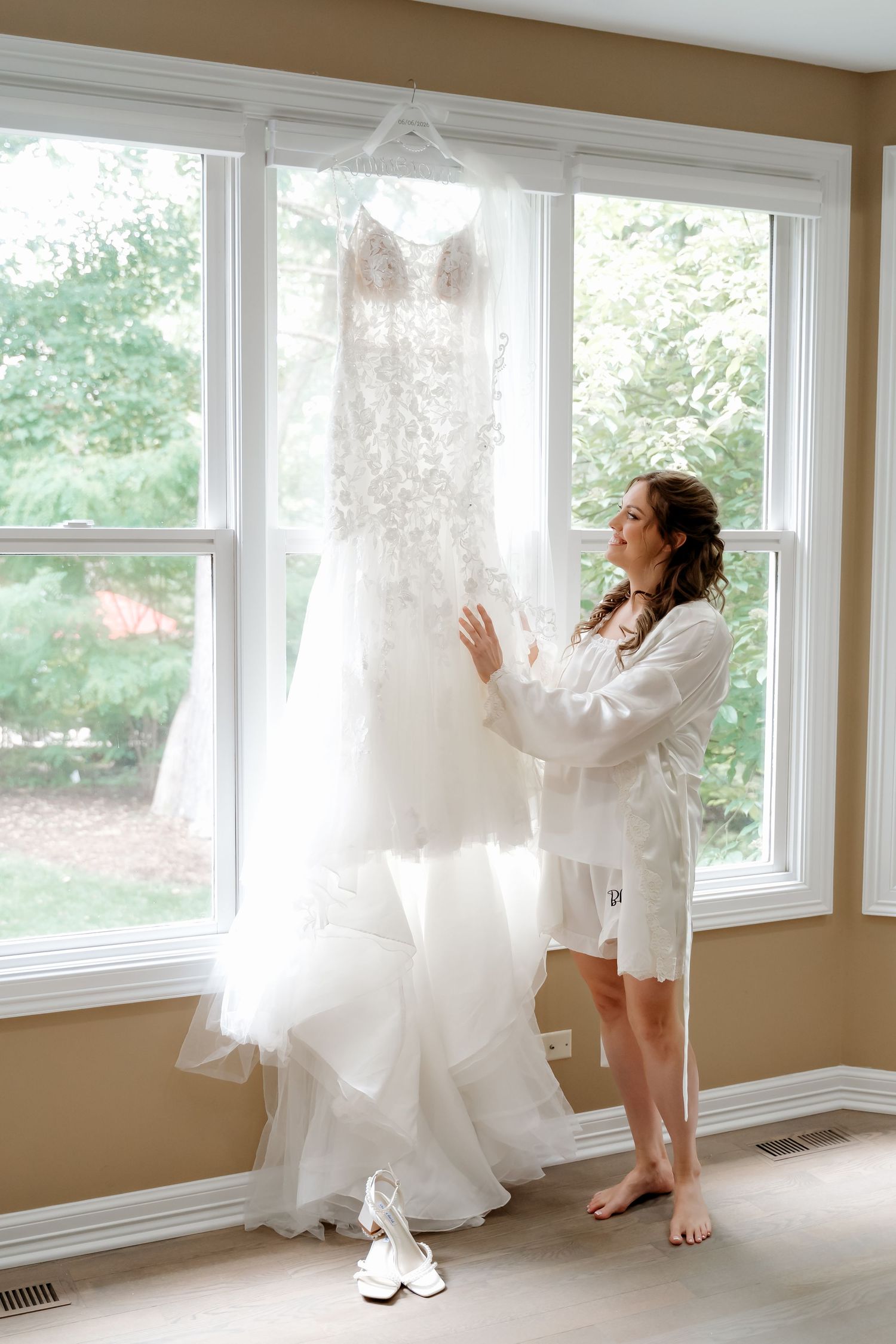 Bride in white robe admiring her lace wedding gown hanging by a bright window with heels on the floor.