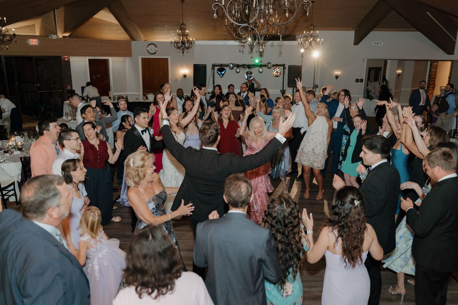 Guests celebrate on the dance floor at a wedding reception in an elegant venue with chandeliers and warm lighting.