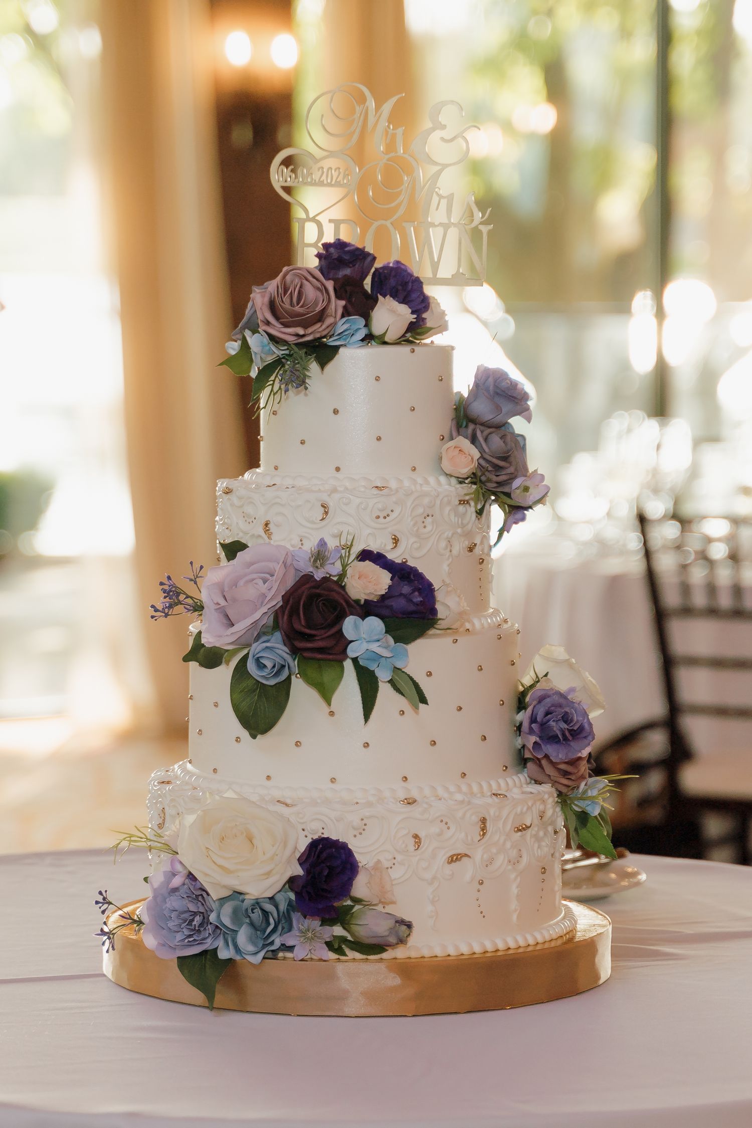 Four-tier white wedding cake with purple and blue floral decorations and Mr & Mrs Brown gold cake topper.