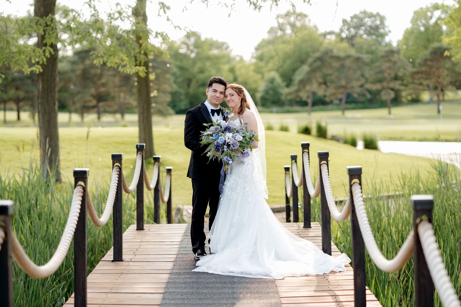 Bride and groom embrace on wooden dock with rope railings, surrounded by greenery, bride holding purple bouquet.