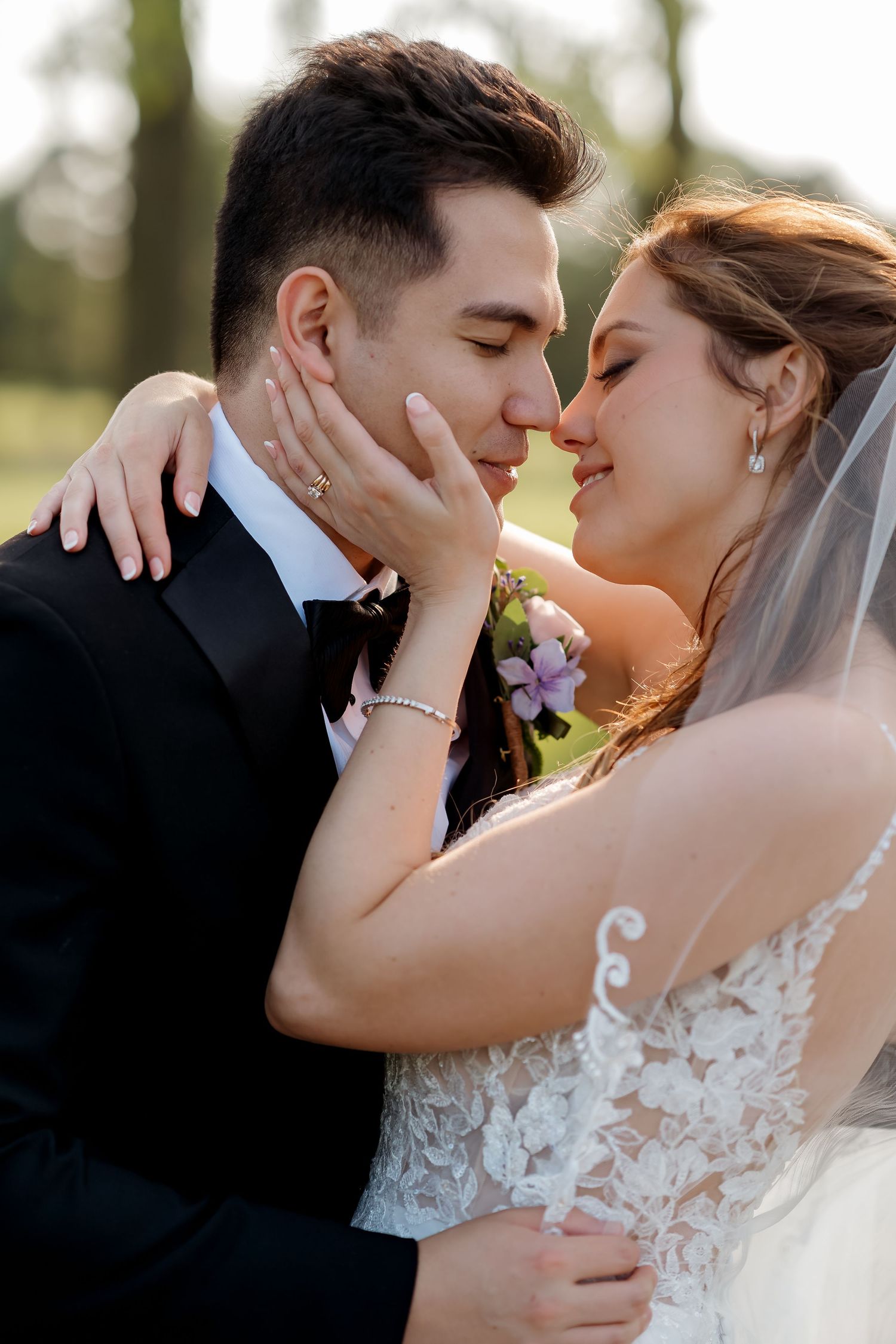 A bride and groom share a romantic almost-kiss moment outdoors, dressed in wedding attire with a floral boutonniere.