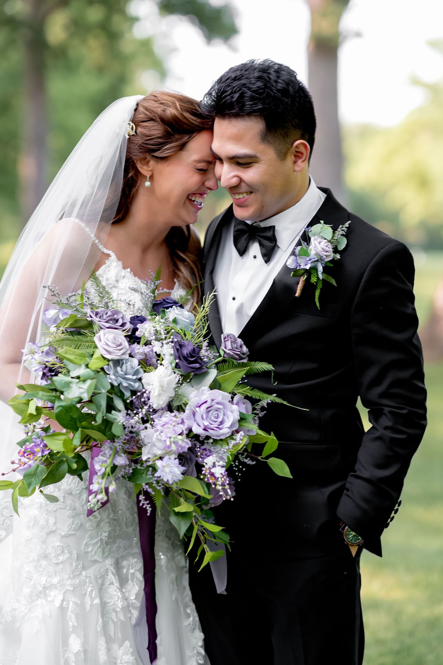 Bride and groom share a joyful moment, touching foreheads, with a lush purple and white floral bouquet outdoors.