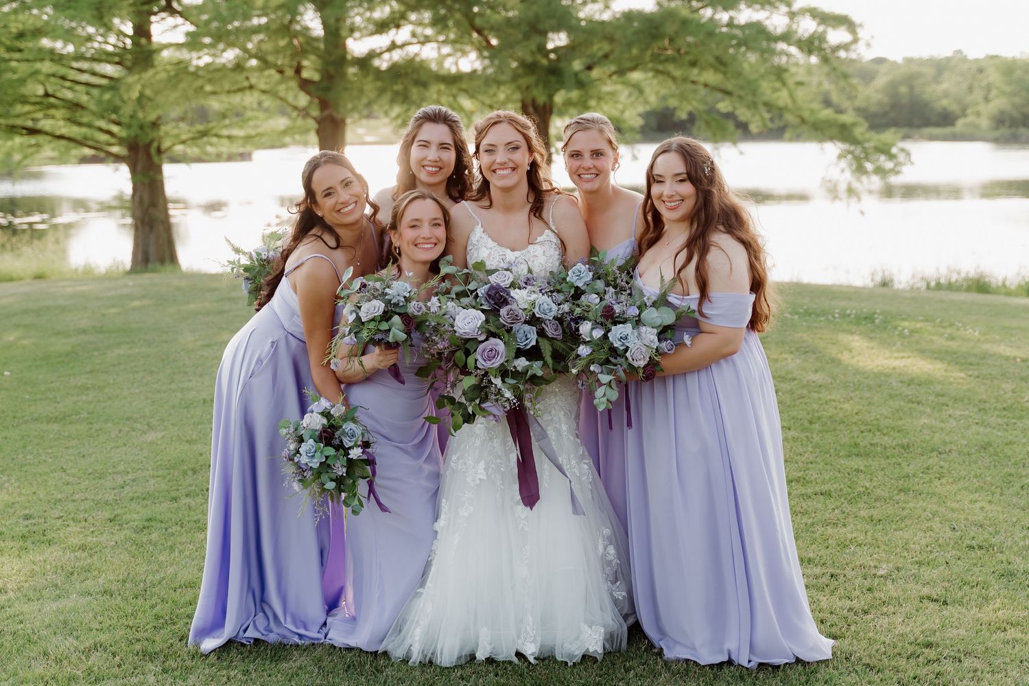 Bride in white gown with five bridesmaids in lavender dresses holding floral bouquets outdoors near a lake.
