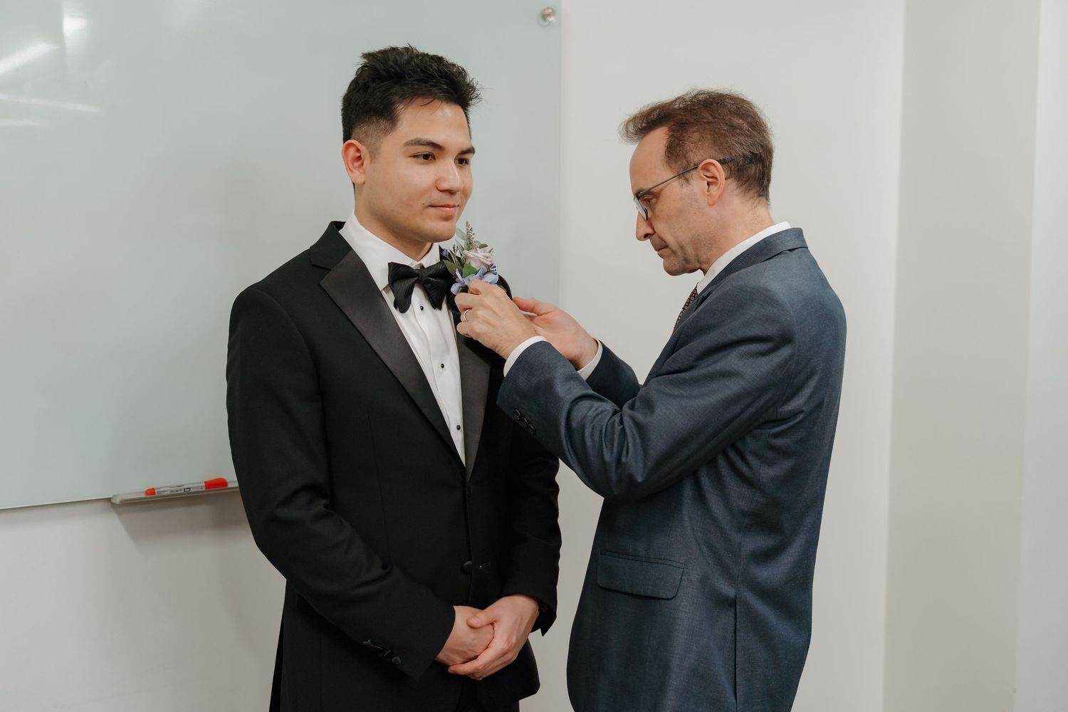 Man in black tuxedo getting boutonniere pinned by older man in gray suit before wedding ceremony.