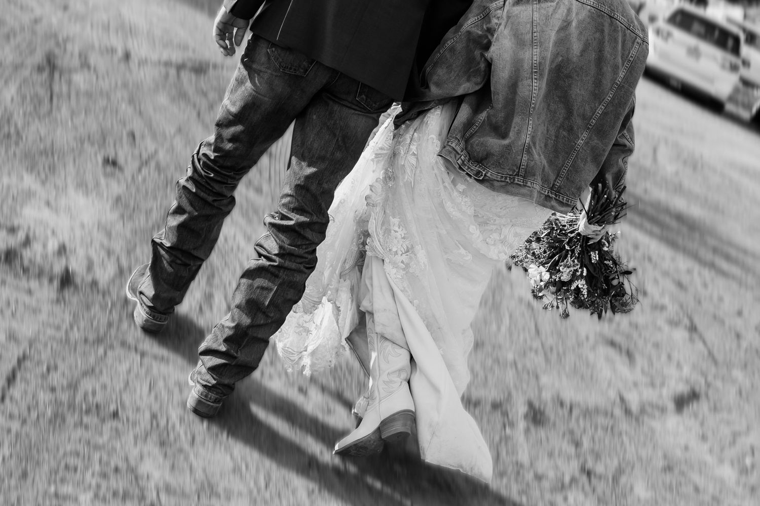 Black and white photo of bride and groom walking, bride in white gown holding bouquet, groom in denim jacket.