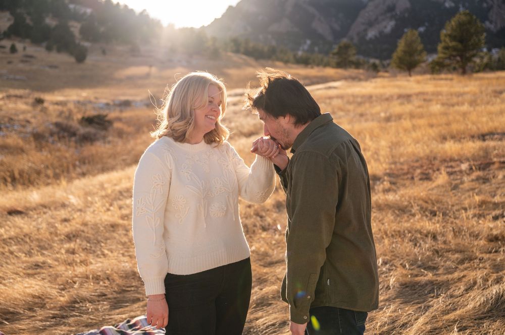 Man kissing woman's hand in golden sunlit field with rocky mountains in background during romantic outdoor photo session.
