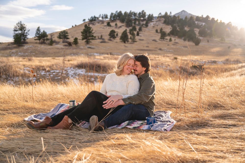 Couple sitting on a blanket in golden meadow with Rocky Mountain foothills backdrop during engagement photo session.