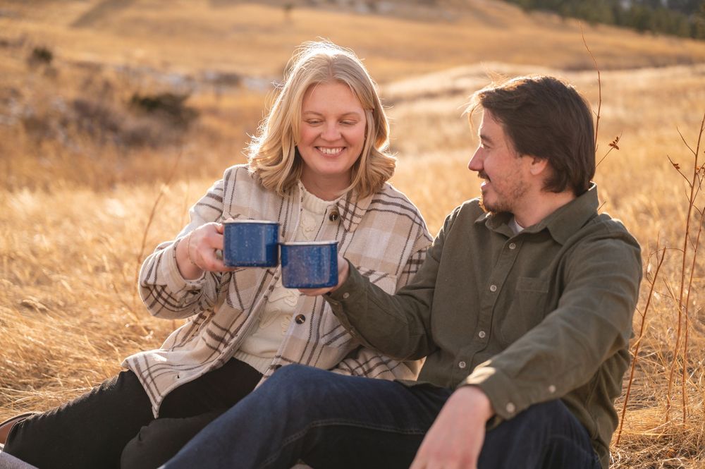 Couple sitting in golden field toasting with blue mugs, smiling warmly on a sunny autumn day outdoors.