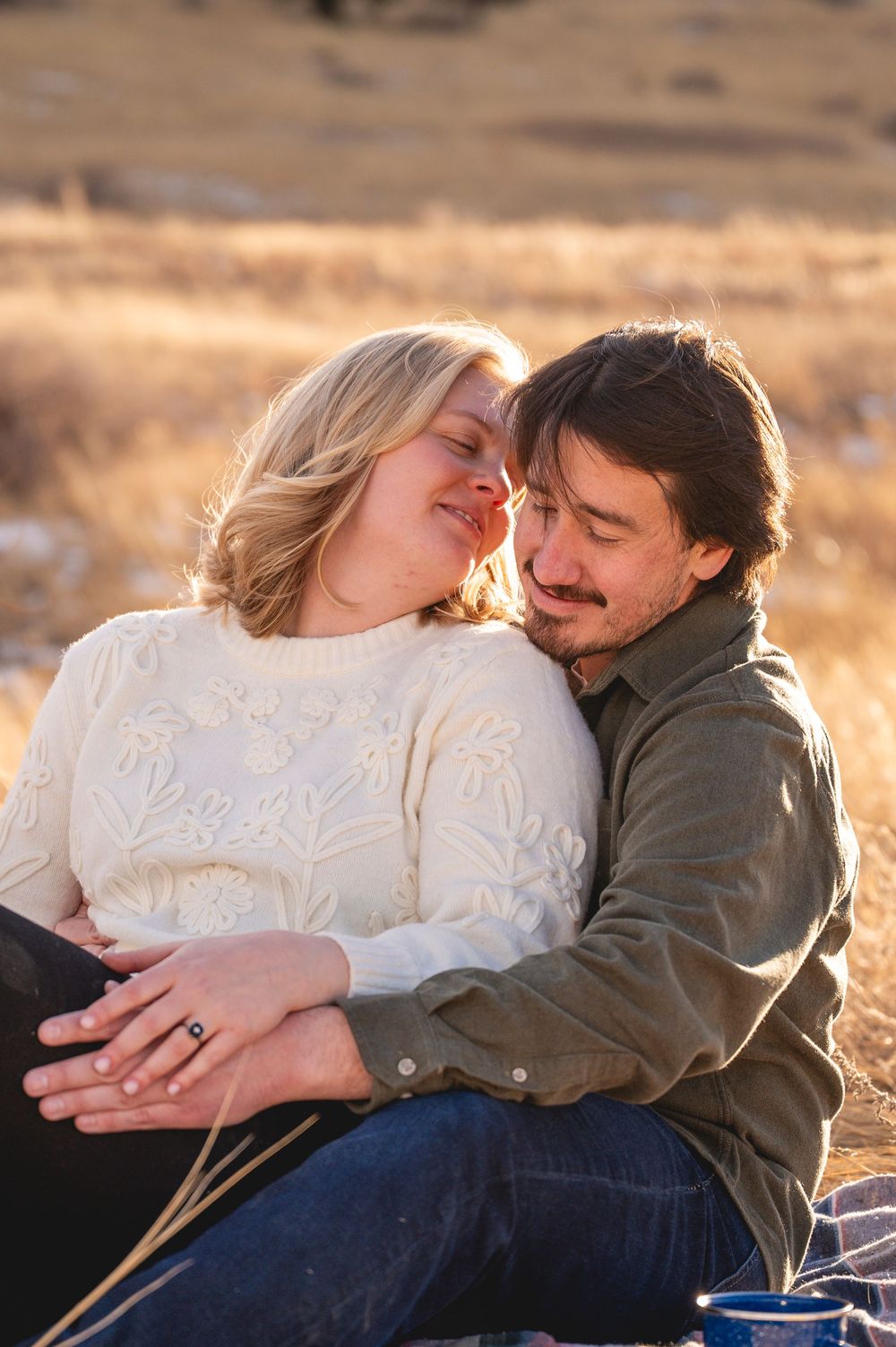 Couple sitting closely together in a golden field, woman in white sweater leaning into man in olive shirt.