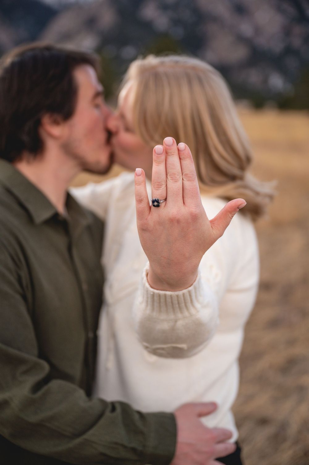 Couple kissing outdoors while showing off engagement ring with dark gemstone, mountains visible in blurred background.