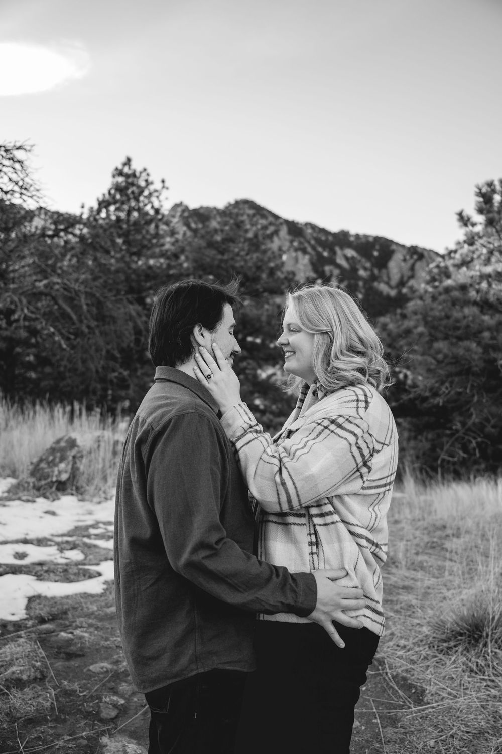 Black and white photo of a couple embracing outdoors, woman touching mans face, mountains and trees in background.