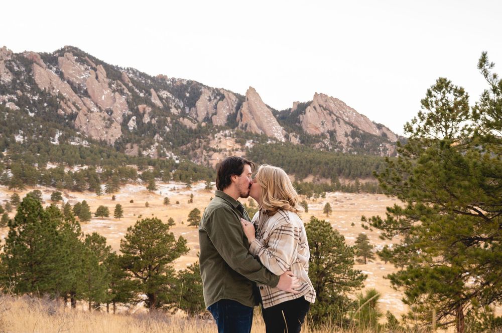 Couple sharing a kiss at Flatirons in Boulder, Colorado, surrounded by pine trees and rocky mountain backdrop.
