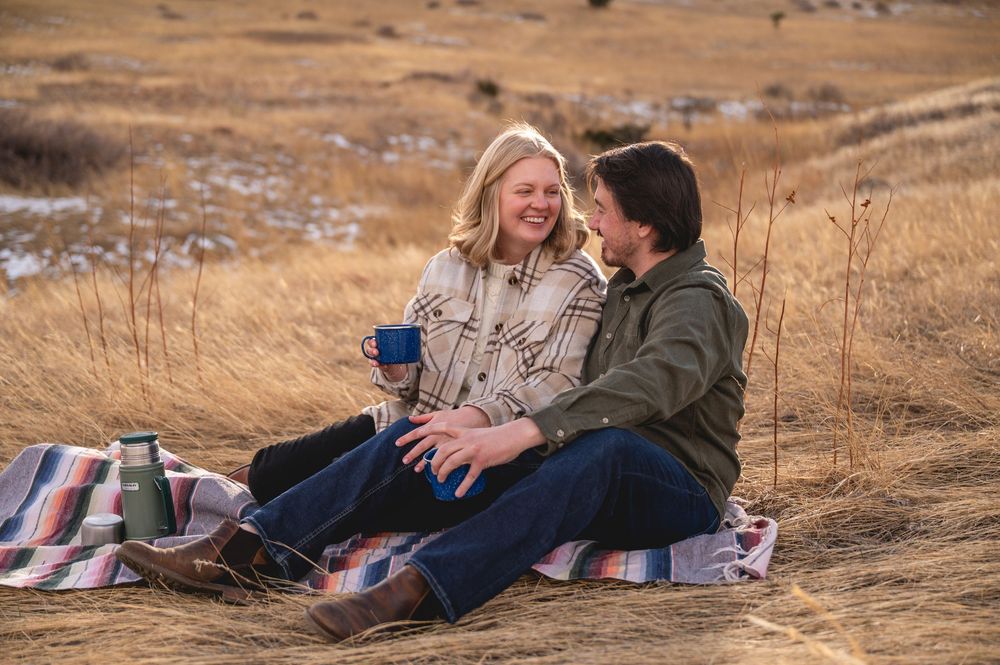 Couple sitting on a blanket in a golden field, sharing warm drinks from blue mugs on a cozy autumn day.