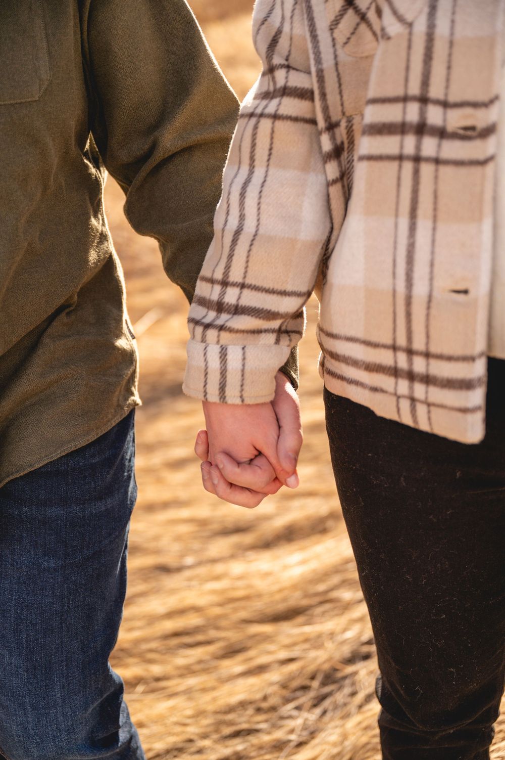 Couple holding hands outdoors in autumn, wearing plaid and olive jackets, with golden dry grass in the background.