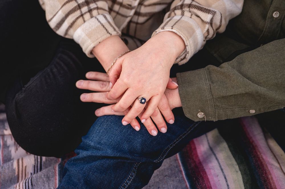 Close-up of two people holding hands showing a black gemstone engagement ring, sitting on a plaid blanket.