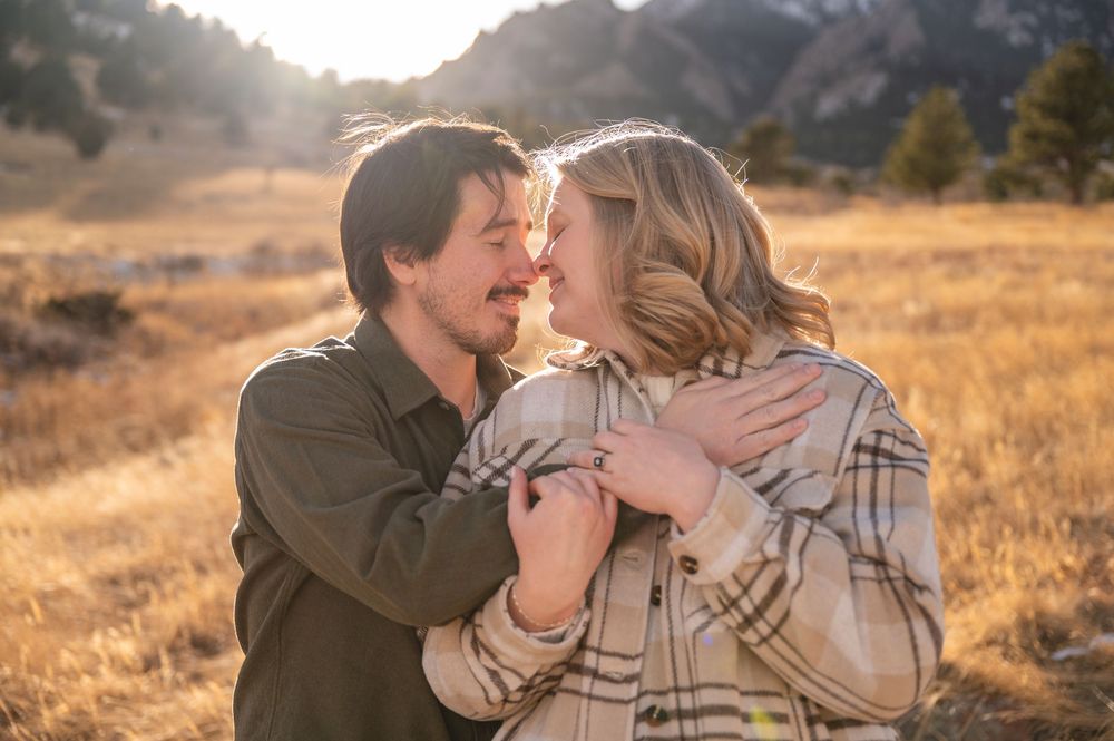 Couple embracing and smiling at each other in a golden sunlit field with mountains in the background.