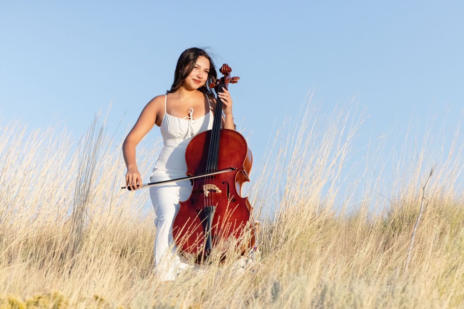 Woman in white dress playing cello outdoors in golden grass field under clear blue sky.