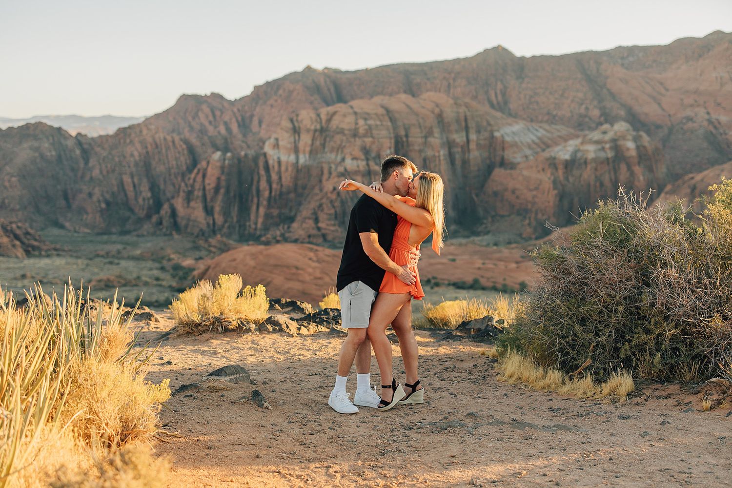 Couple kissing at sunset in a desert landscape with red rock formations in the background.
