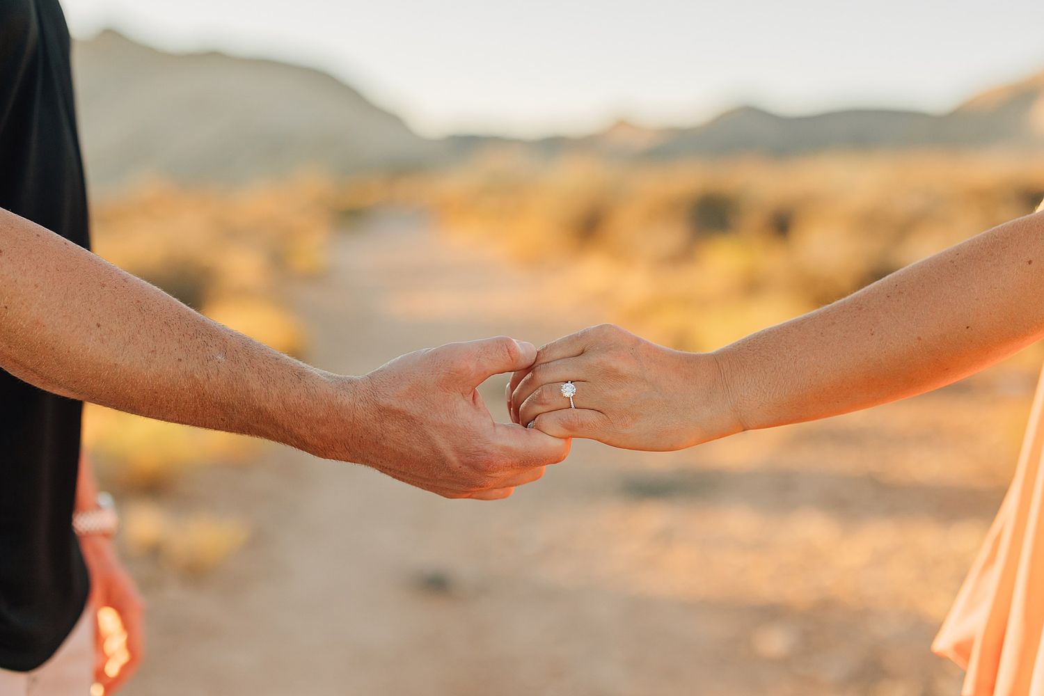 Close-up of couple holding hands showing engagement ring in desert setting.