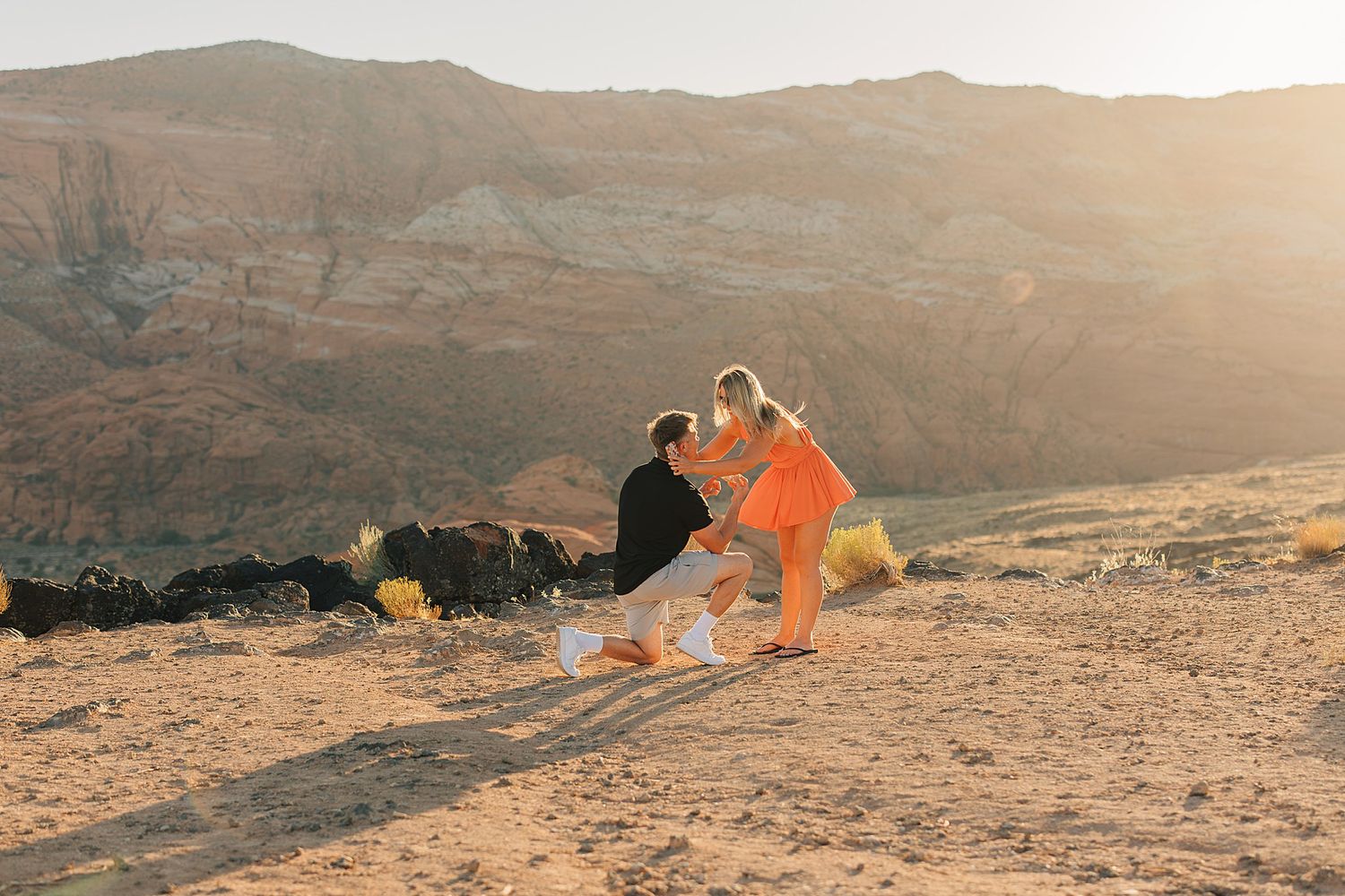 Man proposing to woman in orange dress on desert sand with scenic mountain backdrop at golden hour.