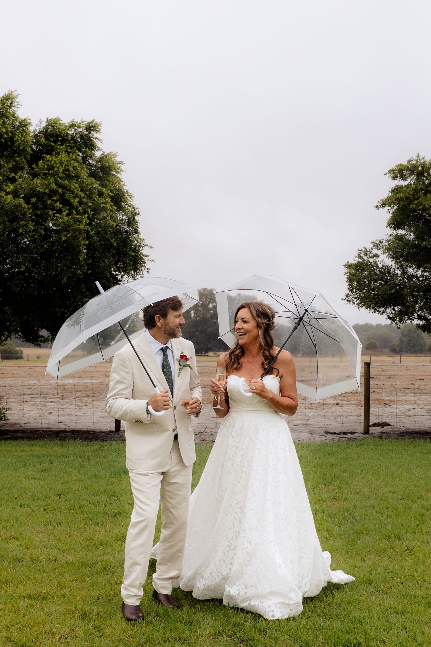 A smiling bride and groom hold clear umbrellas on a rainy wedding day, standing on green grass with trees in the background.