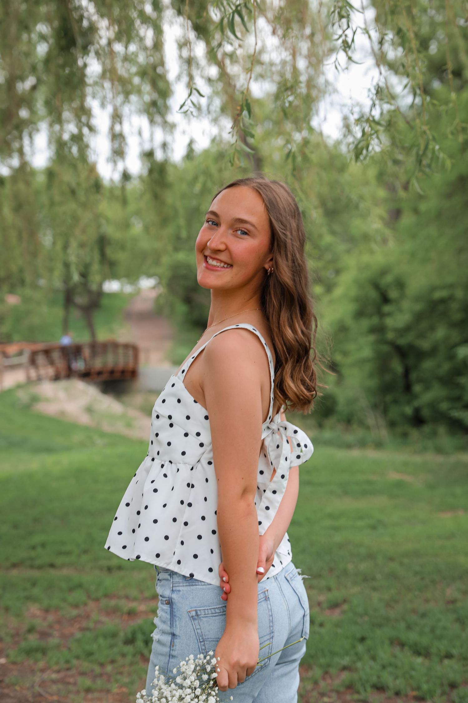 Smiling young woman in polka dot top and jeans holding wildflowers in a lush green park setting.