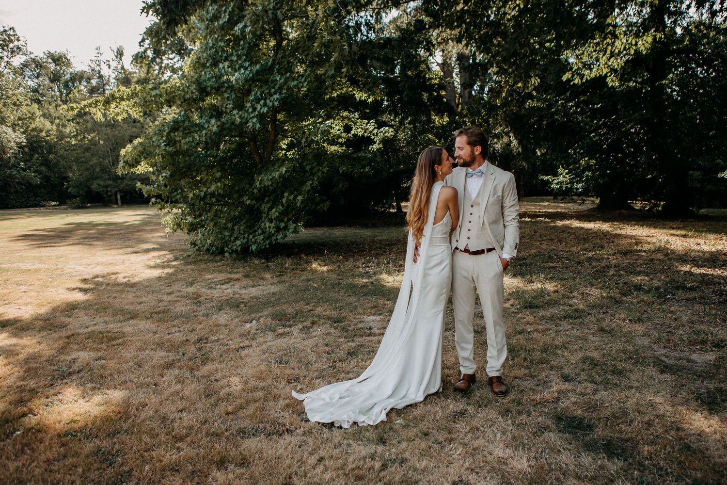 A bride in a flowing white gown and groom in beige suit share a romantic moment in a sunlit park.