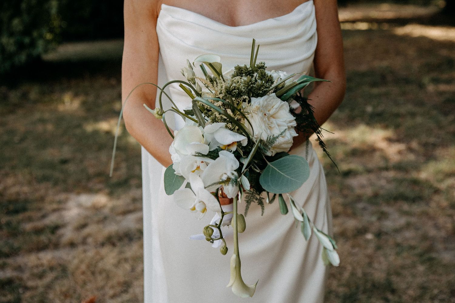 Bride in white gown holding lush cascading wedding bouquet with white flowers and greenery outdoors.