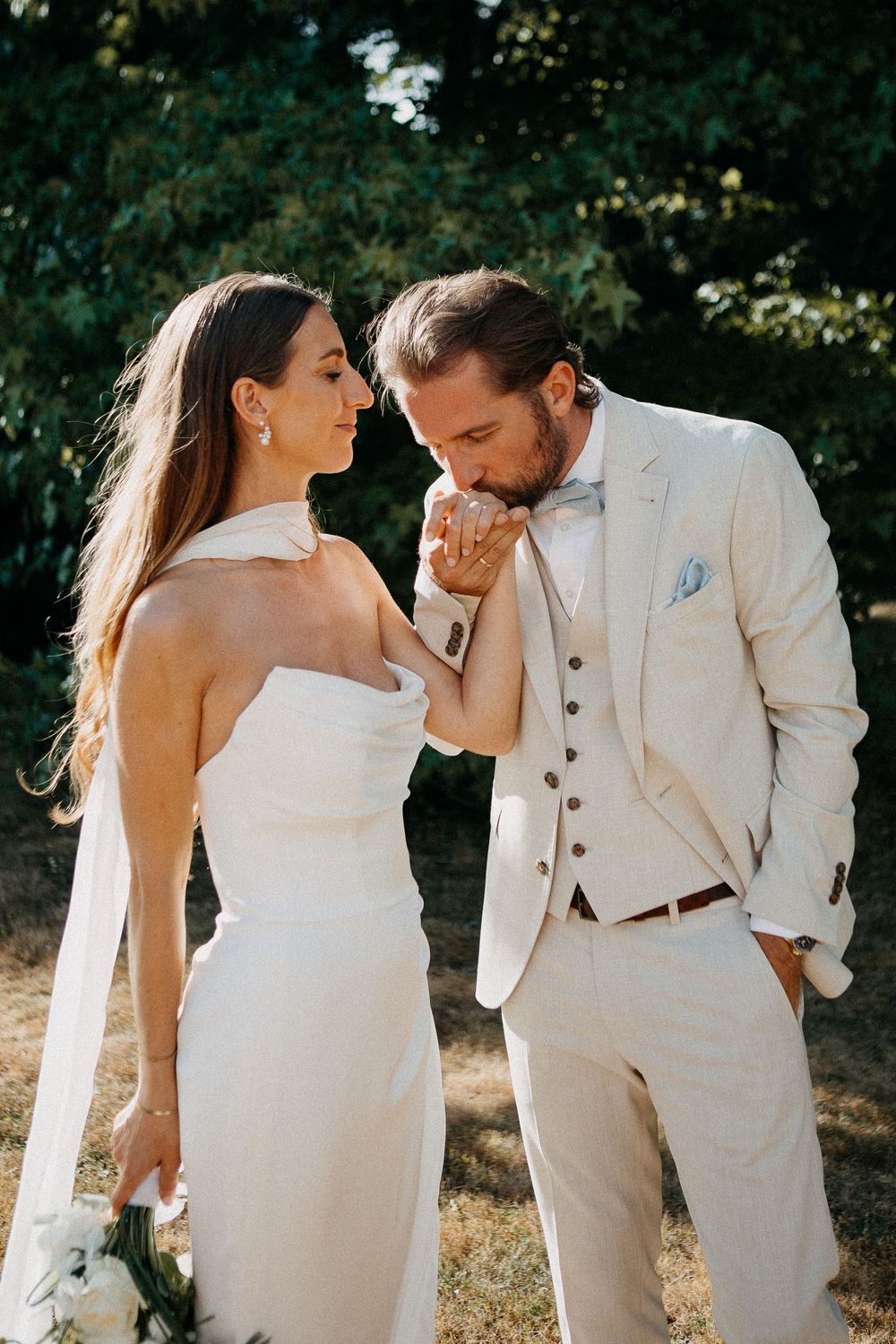 Groom in beige suit kisses bride's hand as she holds white bouquet outdoors.