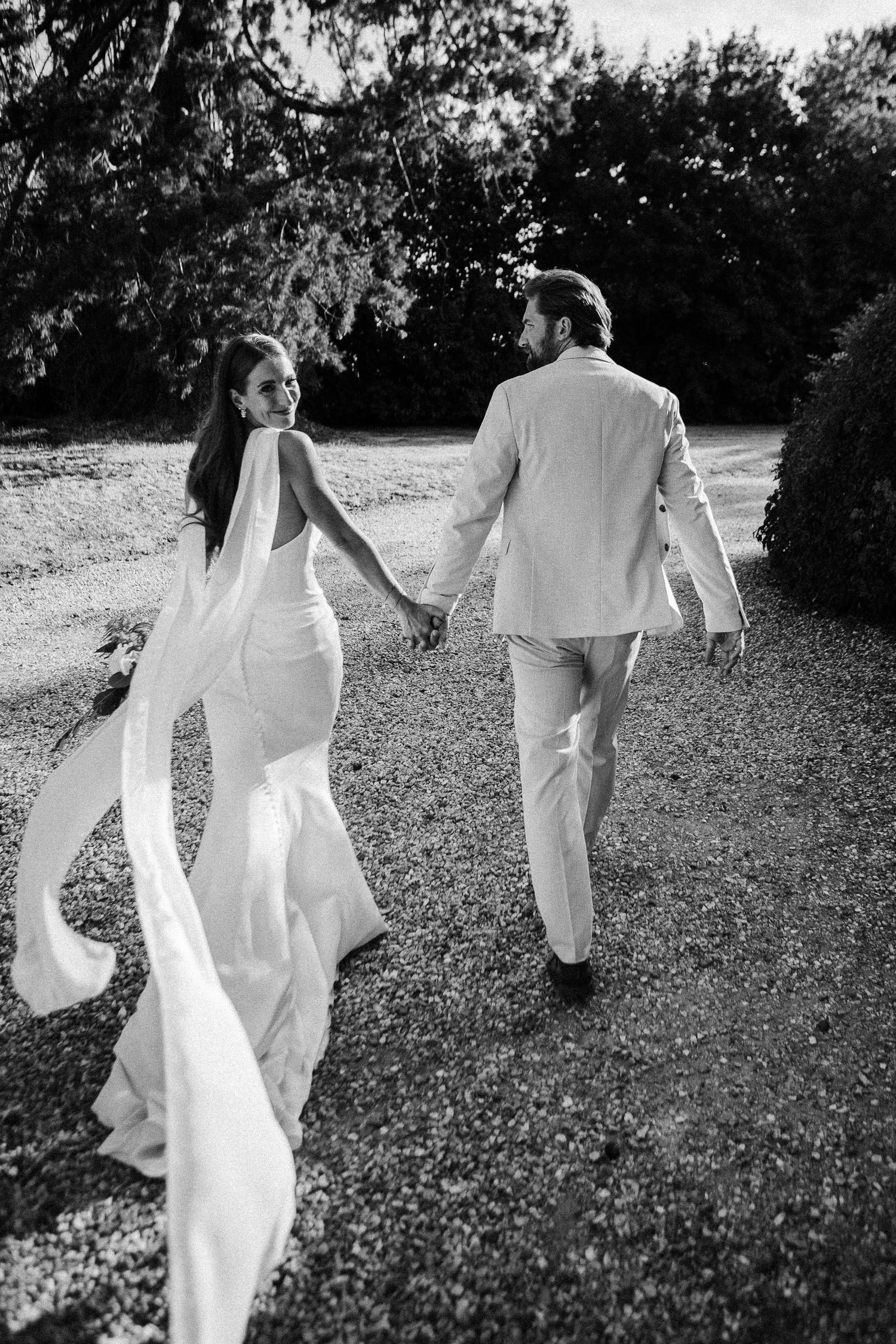 Black and white photo of bride and groom holding hands walking on gravel path.