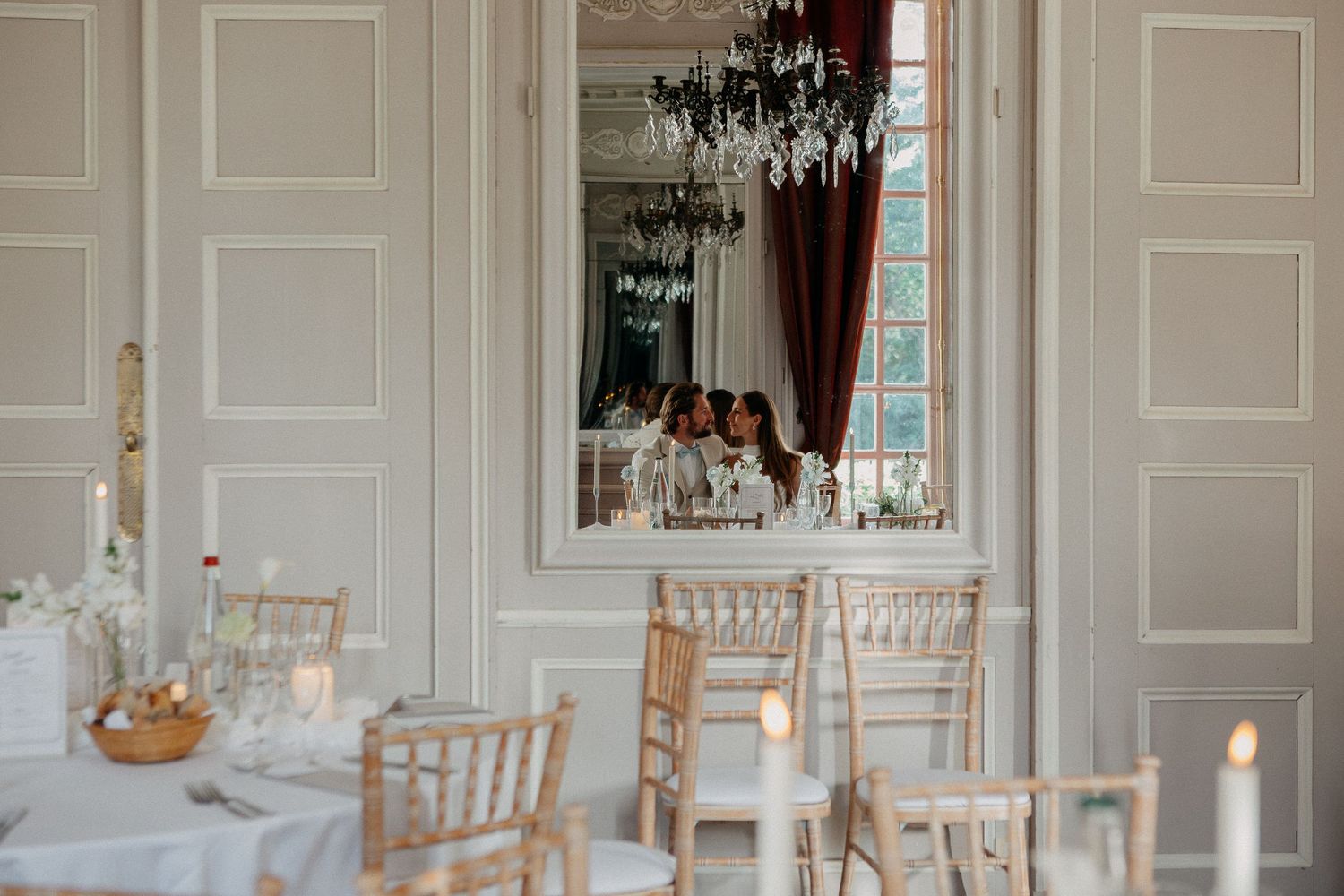 Elegant wedding reception room with chiavari chairs, white linens, and a grand mirror reflecting the bridal party.