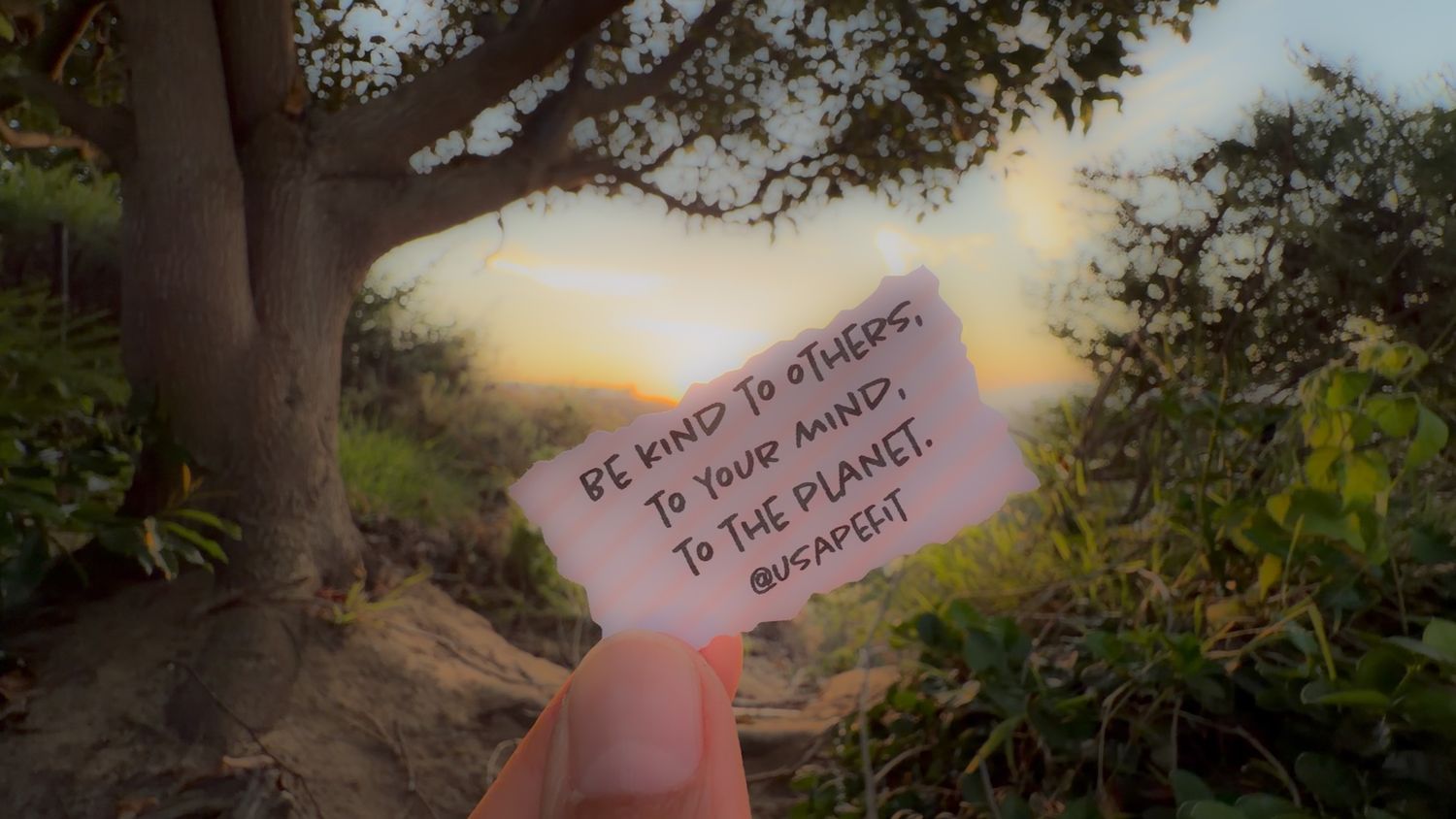 Hand holding a note saying 'Be kind to others, to your mind, to the planet' against a scenic sunset backdrop.