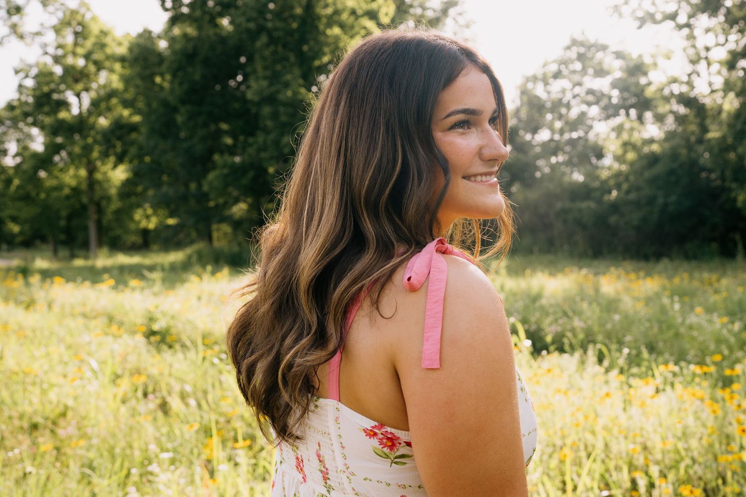 Young woman with long brown hair smiling in a sunny meadow, wearing a pink floral dress with a shoulder tattoo.