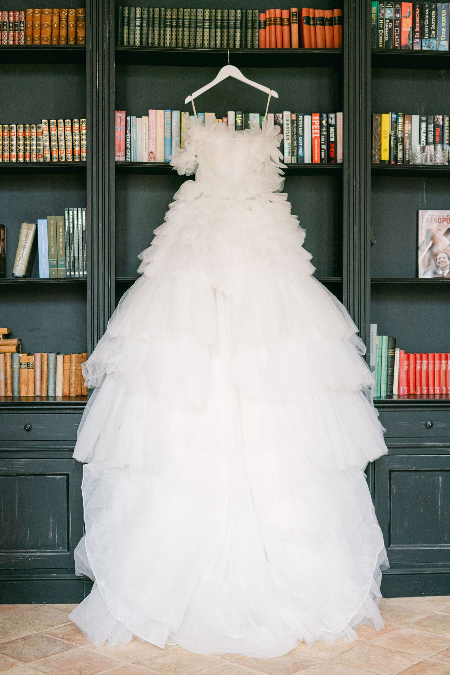 Elegant white tiered tulle ball gown wedding dress hanging on a hanger in front of dark green bookshelves.