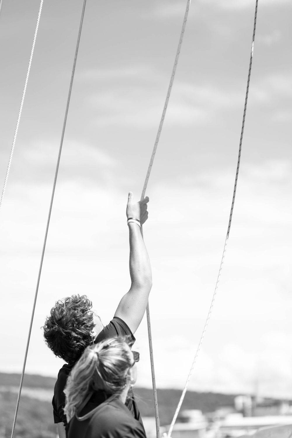 Black and white photo of a person reaching up to grab sailing ropes on a boat against a cloudy sky.