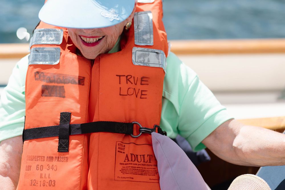 Person wearing an orange adult life vest labeled True Love on a boat, smiling and wearing a white cap.
