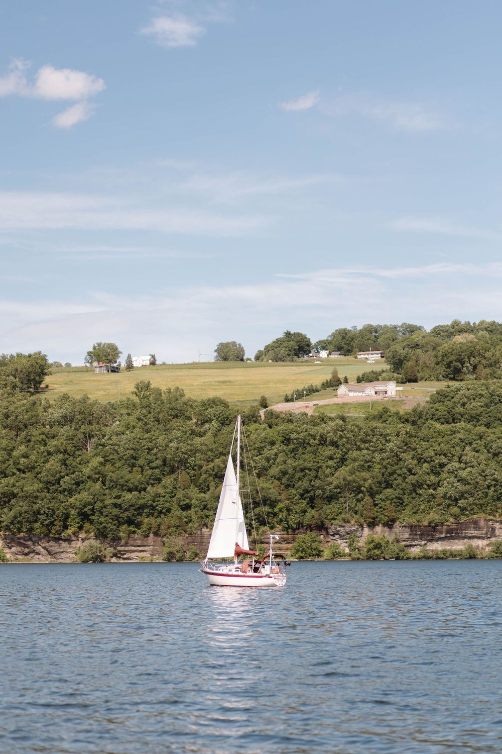 A white sailboat glides across a calm blue lake with lush green tree-covered hills and a partly cloudy sky.