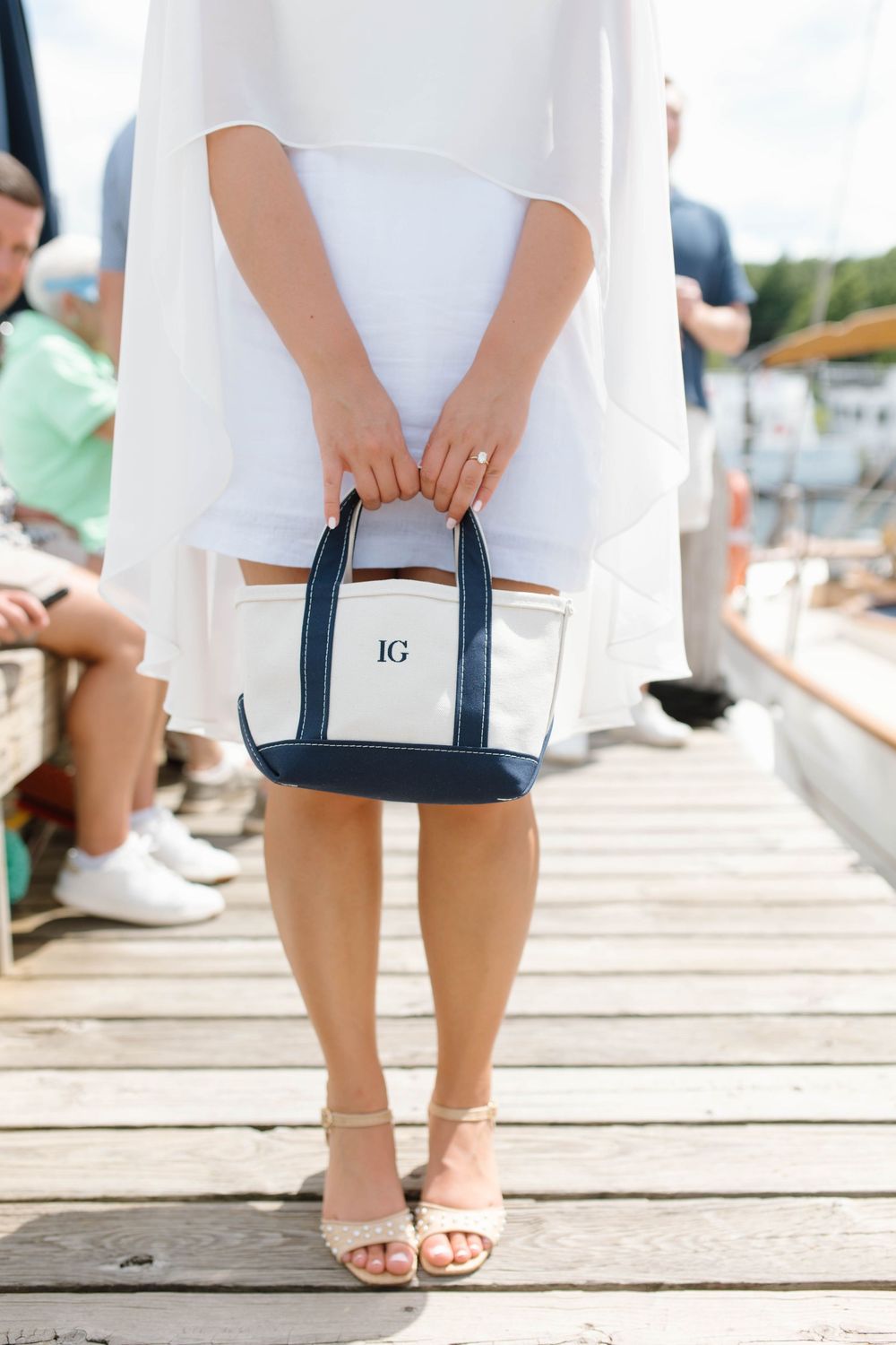 Woman in white dress holding a monogrammed navy and white canvas tote bag on a wooden dock near sailboats.