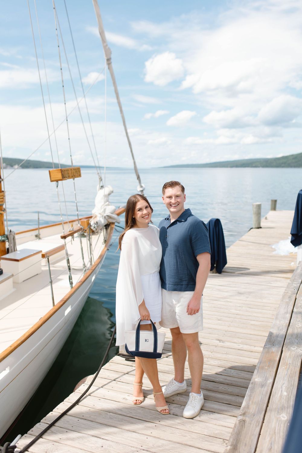 Smiling couple posing on a wooden dock beside a classic sailboat on a beautiful sunny day.