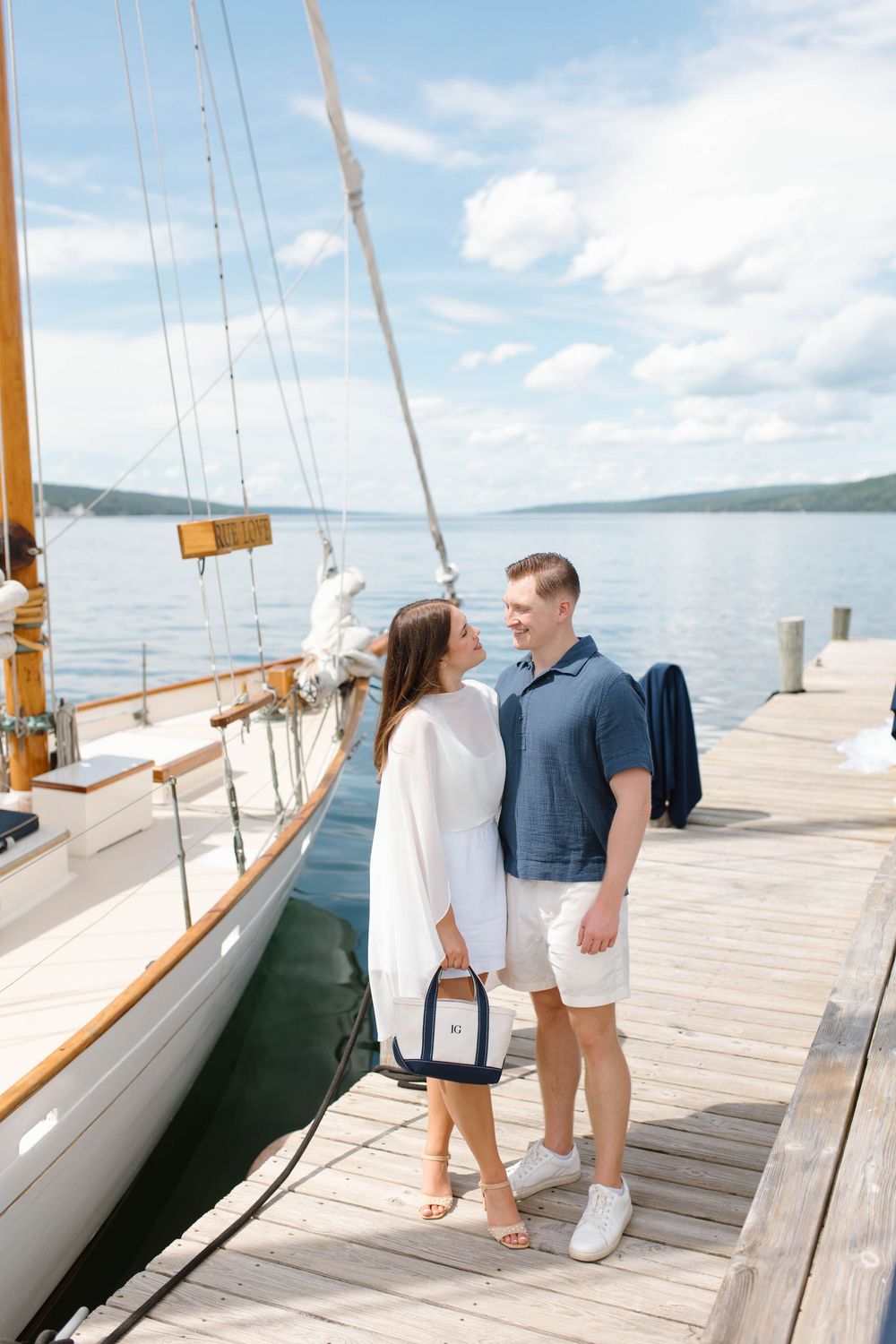 Couple smiling at each other on a sunny dock beside a classic wooden sailboat on calm blue water.