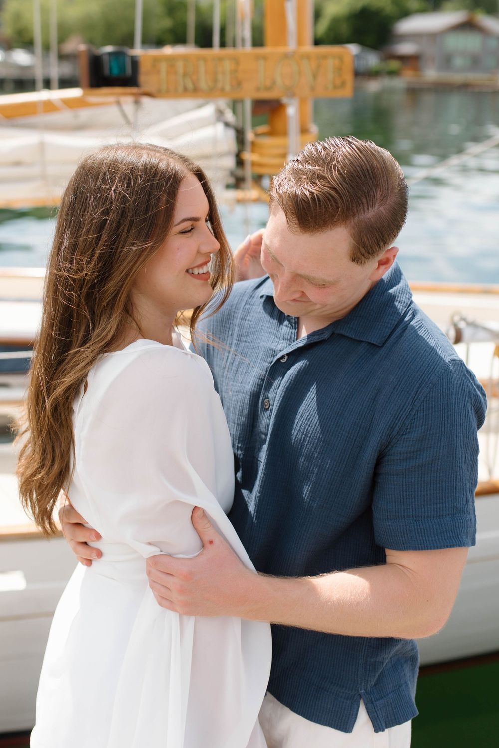 Couple embracing and smiling near a sailboat with a True Love sign at a sunny marina.