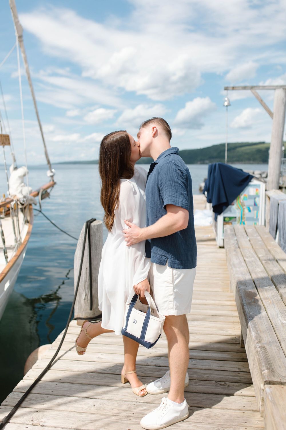 Couple sharing a romantic kiss on a wooden marina dock with sailboats and blue water in the background.